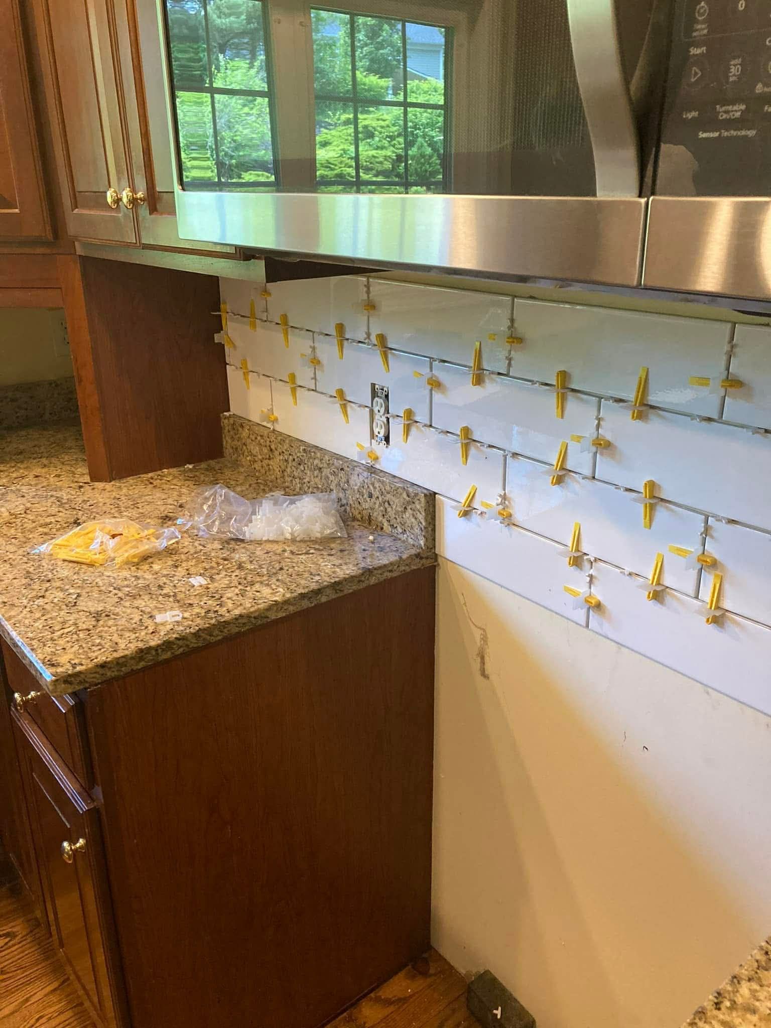 Kitchen backsplash tile installation in progress. White tiles with gold spacers, stainless steel hood.