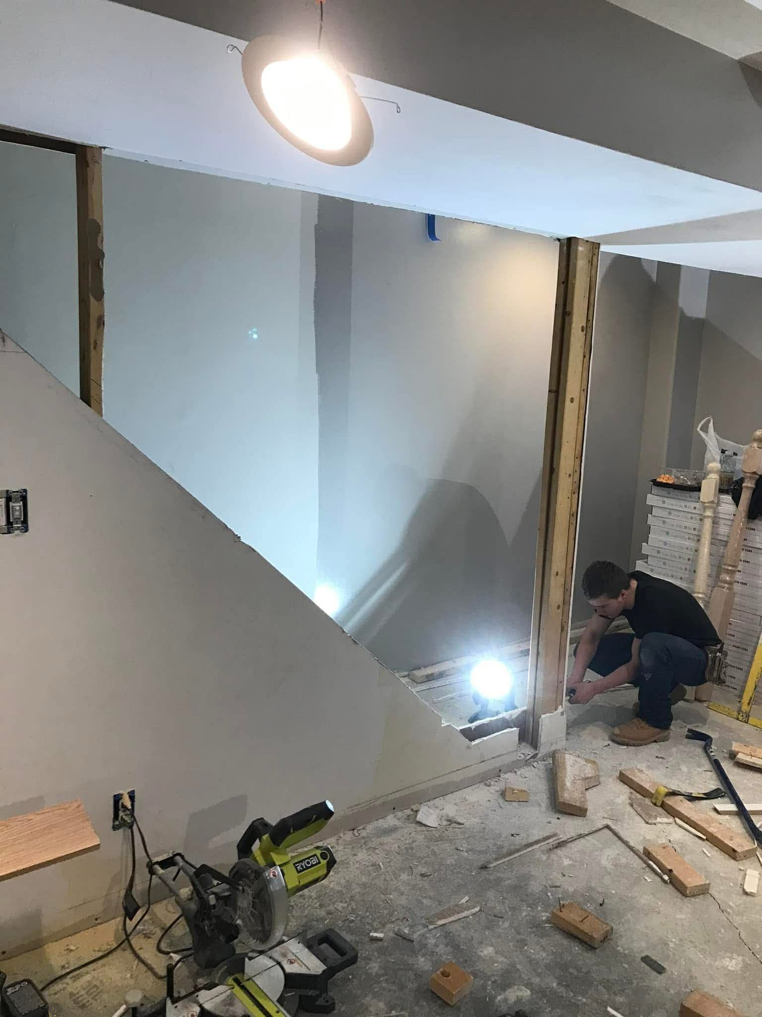 Construction worker renovating a staircase. Drywall, tools, and framing visible. Bright overhead light.