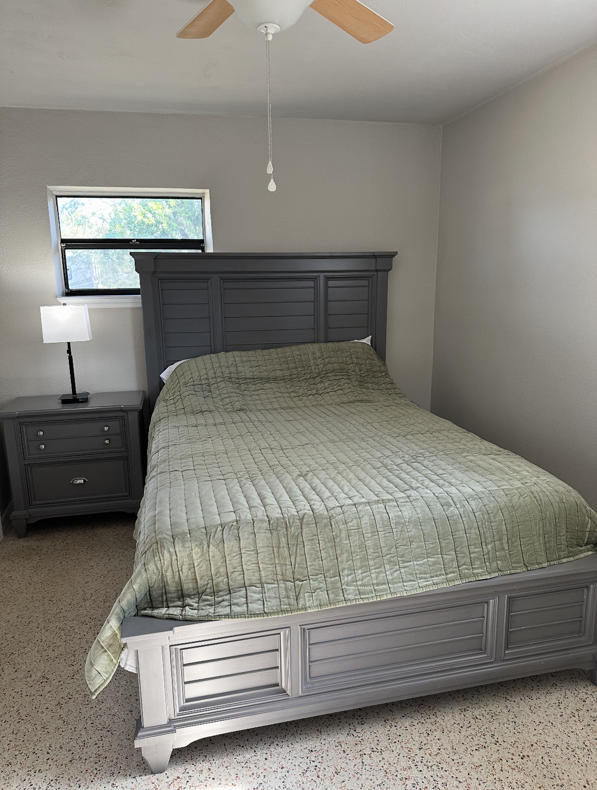 A gray bedroom set with a shutter-style headboard, a matching nightstand with a lamp, and a green textured quilt.