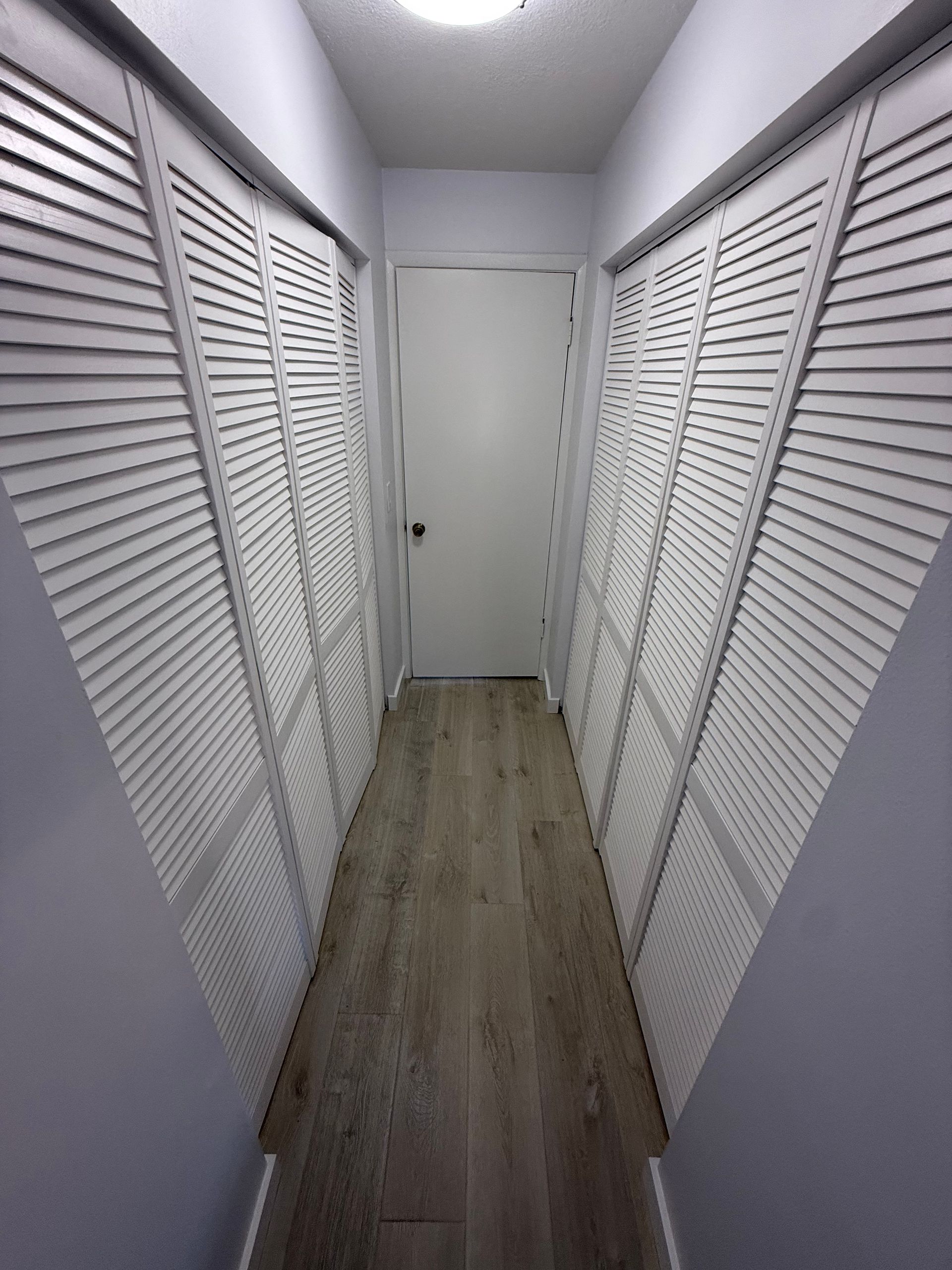 A narrow hallway with white louvered closet doors on both sides, light wood-look flooring, and a door at the end.