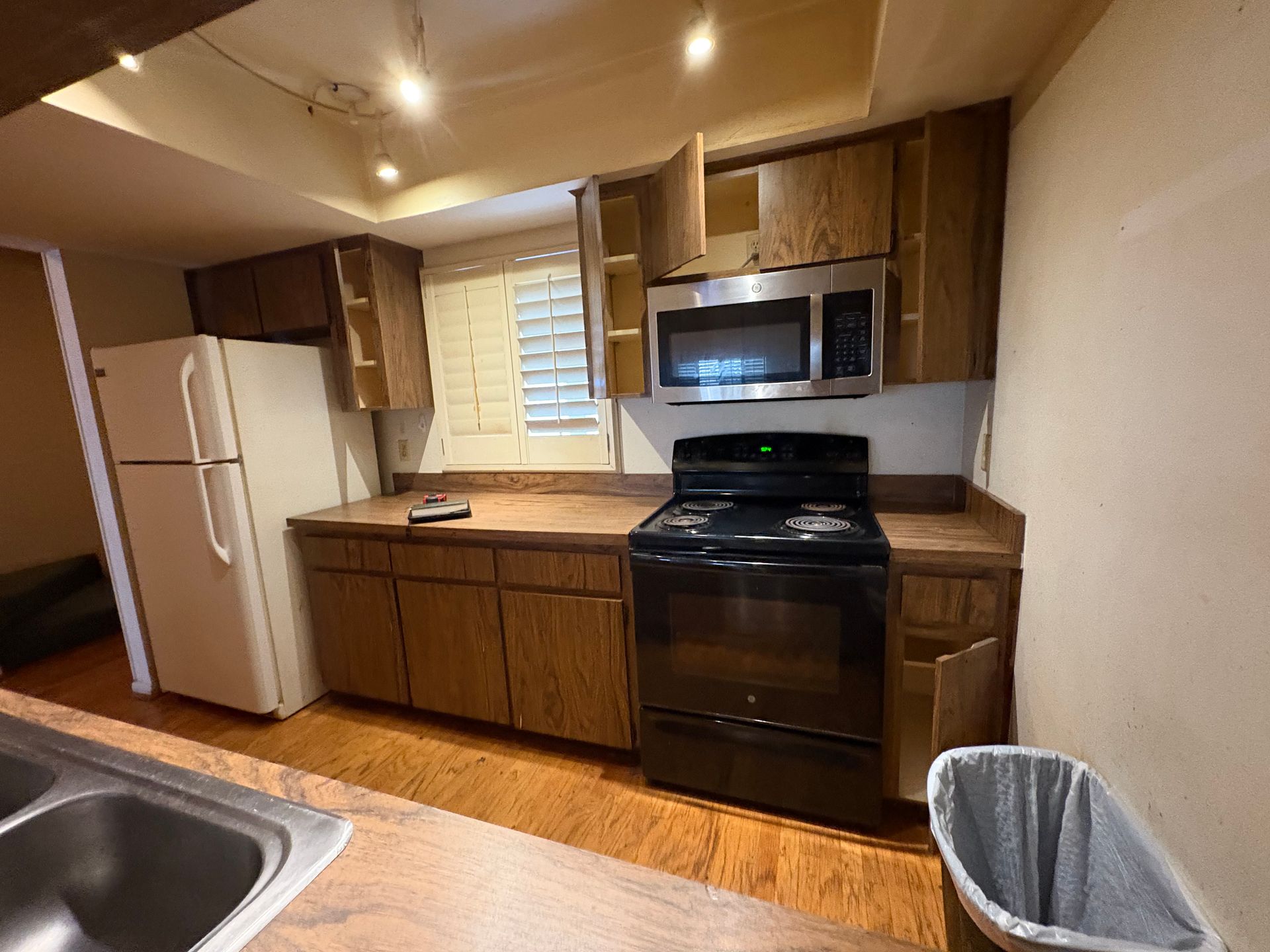 Kitchen with wood cabinets, stainless steel microwave, black stove, white refrigerator, and a trash can on a wood floor.