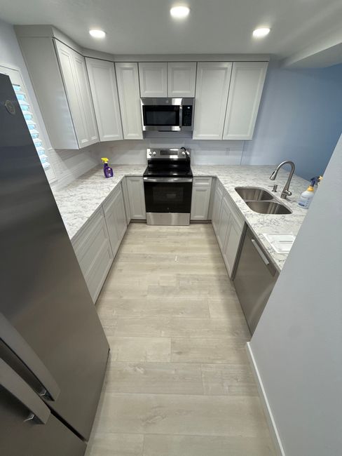 Modern galley kitchen with white cabinets, speckled countertops, stainless steel appliances, and light wood-look flooring.