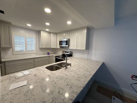 A modern kitchen with grey cabinets, white marble-patterned countertops, a central sink, and recessed ceiling lighting.