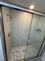 A walk-in shower with a glass door, light grey tile walls, a pebble-stone floor, and a shower head.