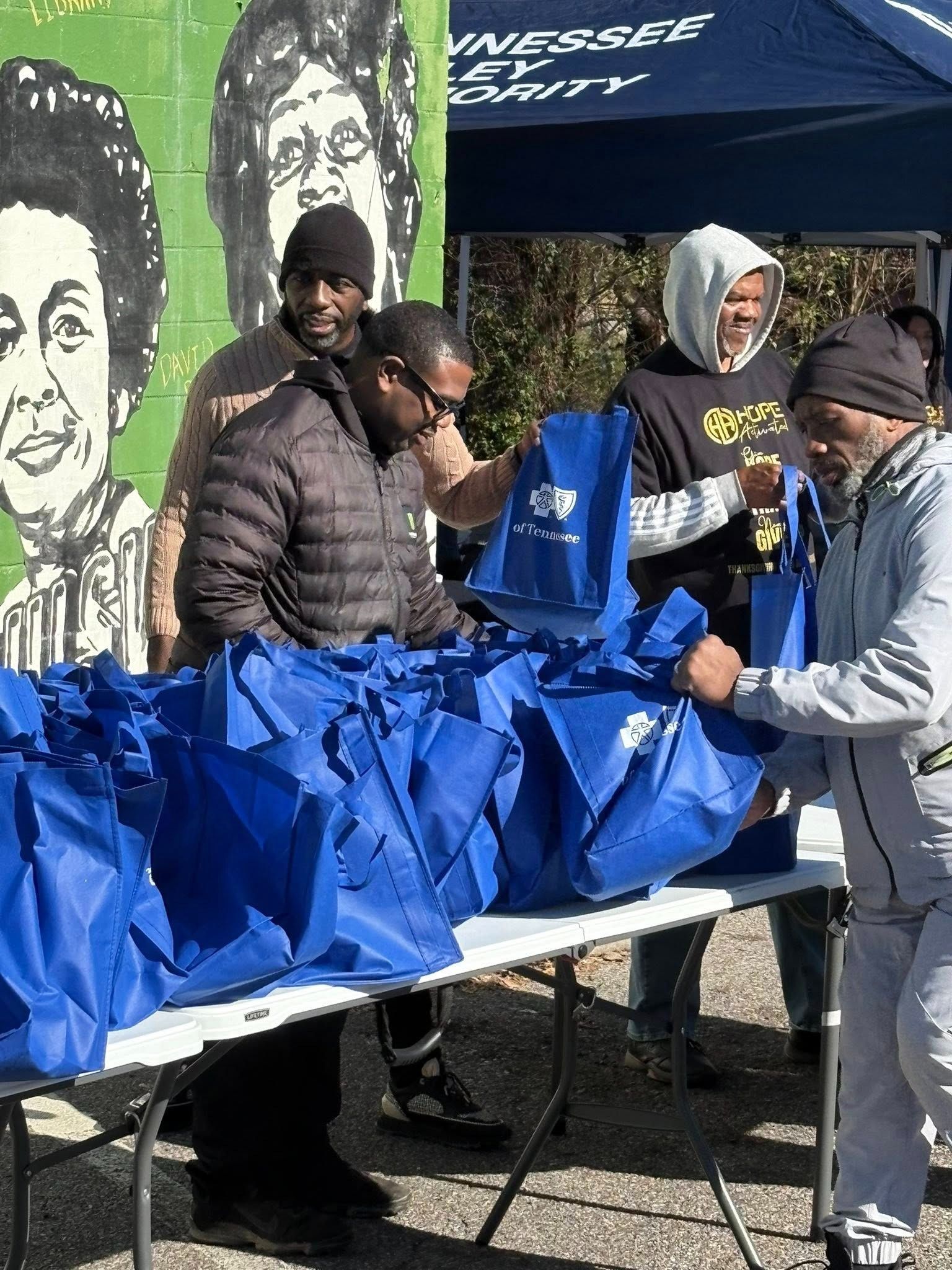 Four people at a table filling blue bags. Mural of women in the background. Outdoor setting.