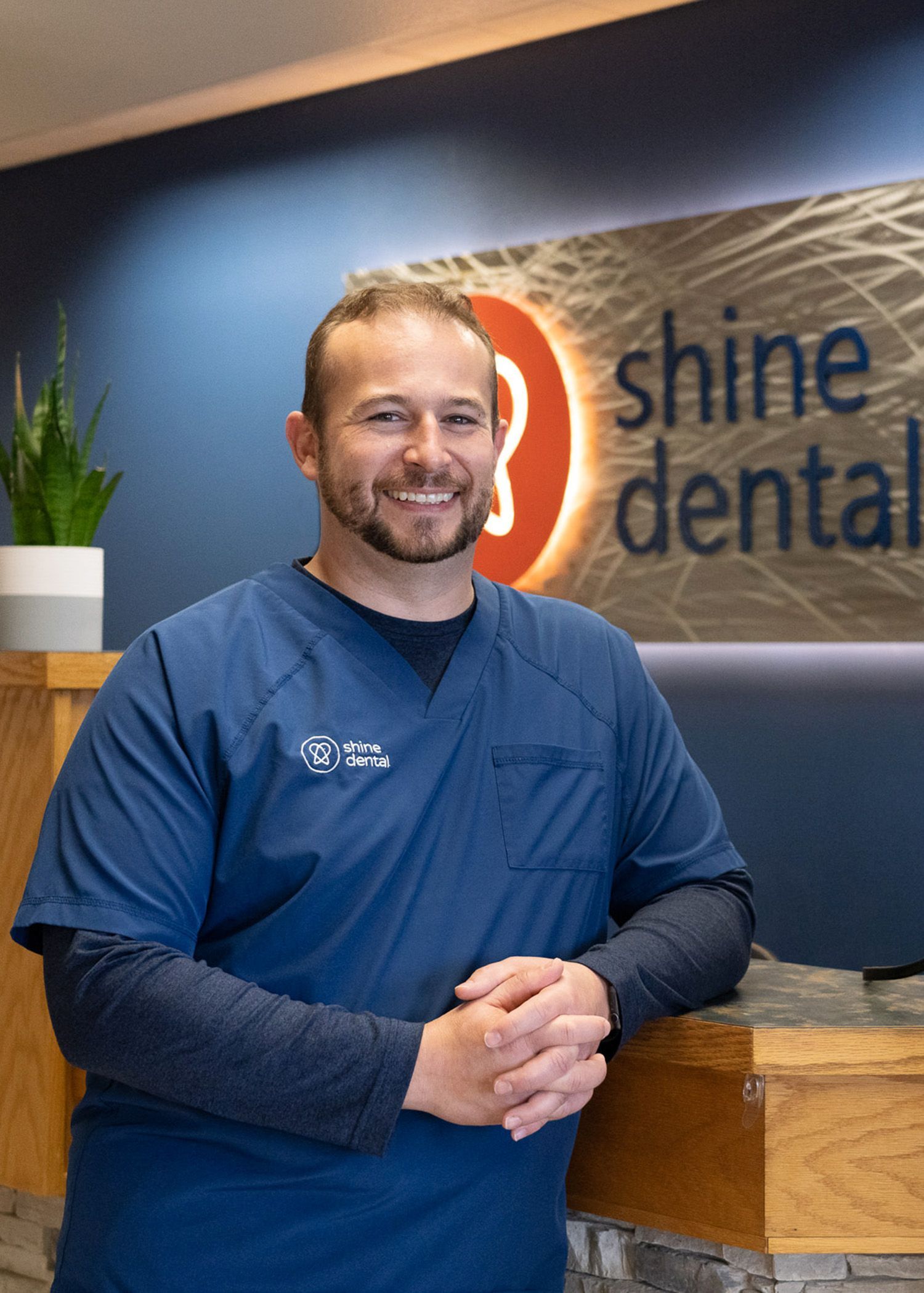 Dr. J in a blue scrub is standing in front of a shine dental sign.
