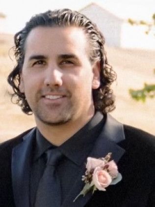 Man in a black suit with a pink boutonniere, smiling outdoors at a wedding