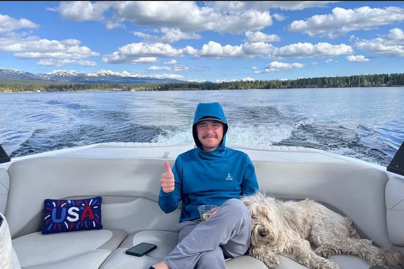 Person in blue jacket and hat giving a thumbs-up on a boat with a dog, lake and mountains behind.