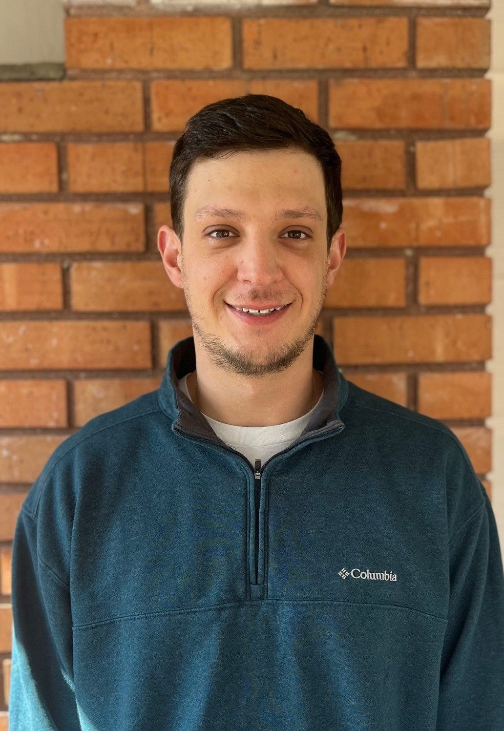 Person in a teal pullover smiling in front of a brick wall.