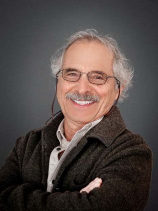 Smiling man with gray hair and glasses, arms crossed against a dark background