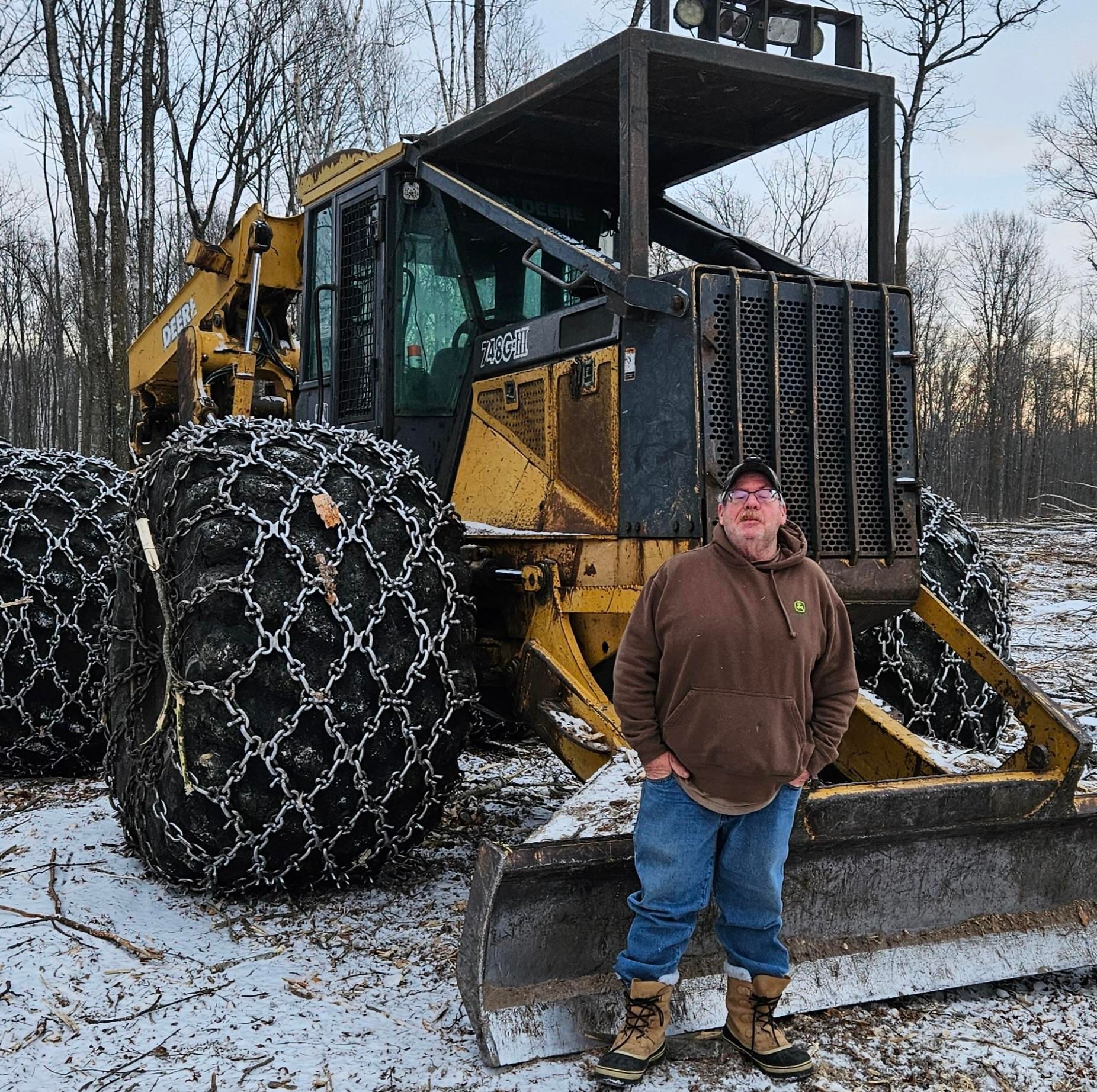 Man standing beside a yellow log skidder with large chain-wrapped tires in a snowy лес clearing