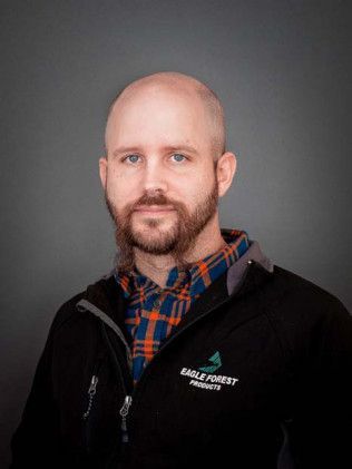 Portrait of a bald man with a beard wearing a black jacket over a plaid shirt against a gray background
