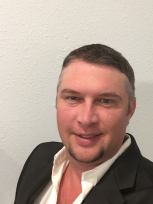 Smiling person in a black blazer and open-collar white shirt against a light wall