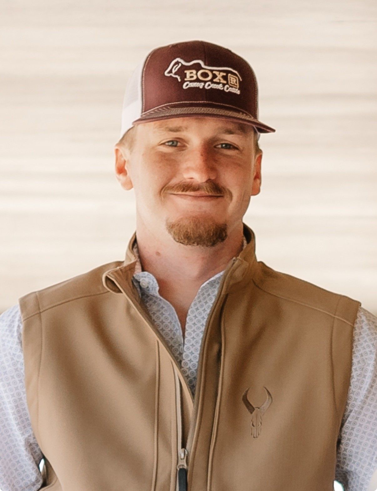 Smiling person in a brown cap and tan vest standing against a light background