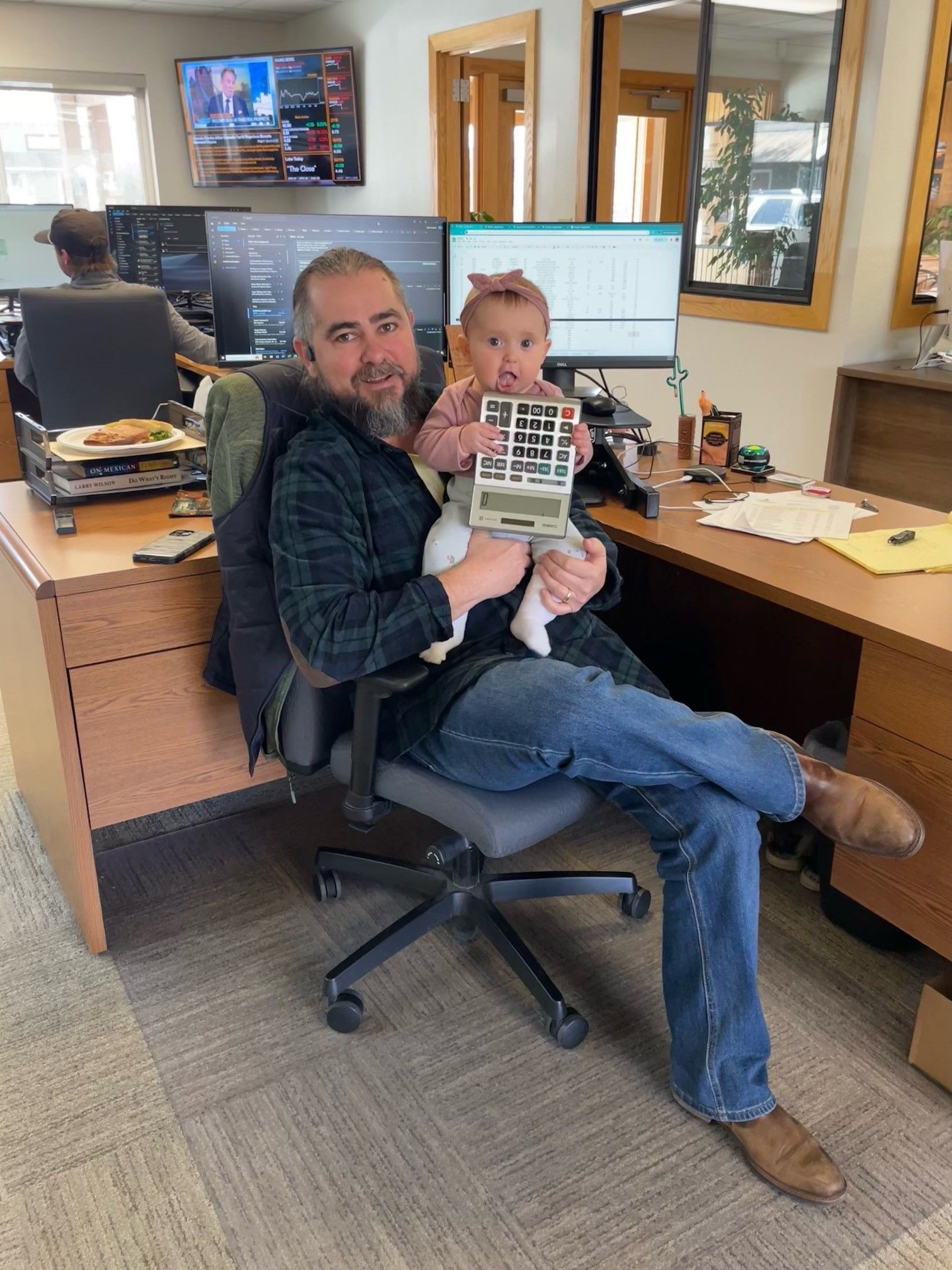 Man holding a toddler in an office chair in a workspace, with desks, computers, and wall art behind them