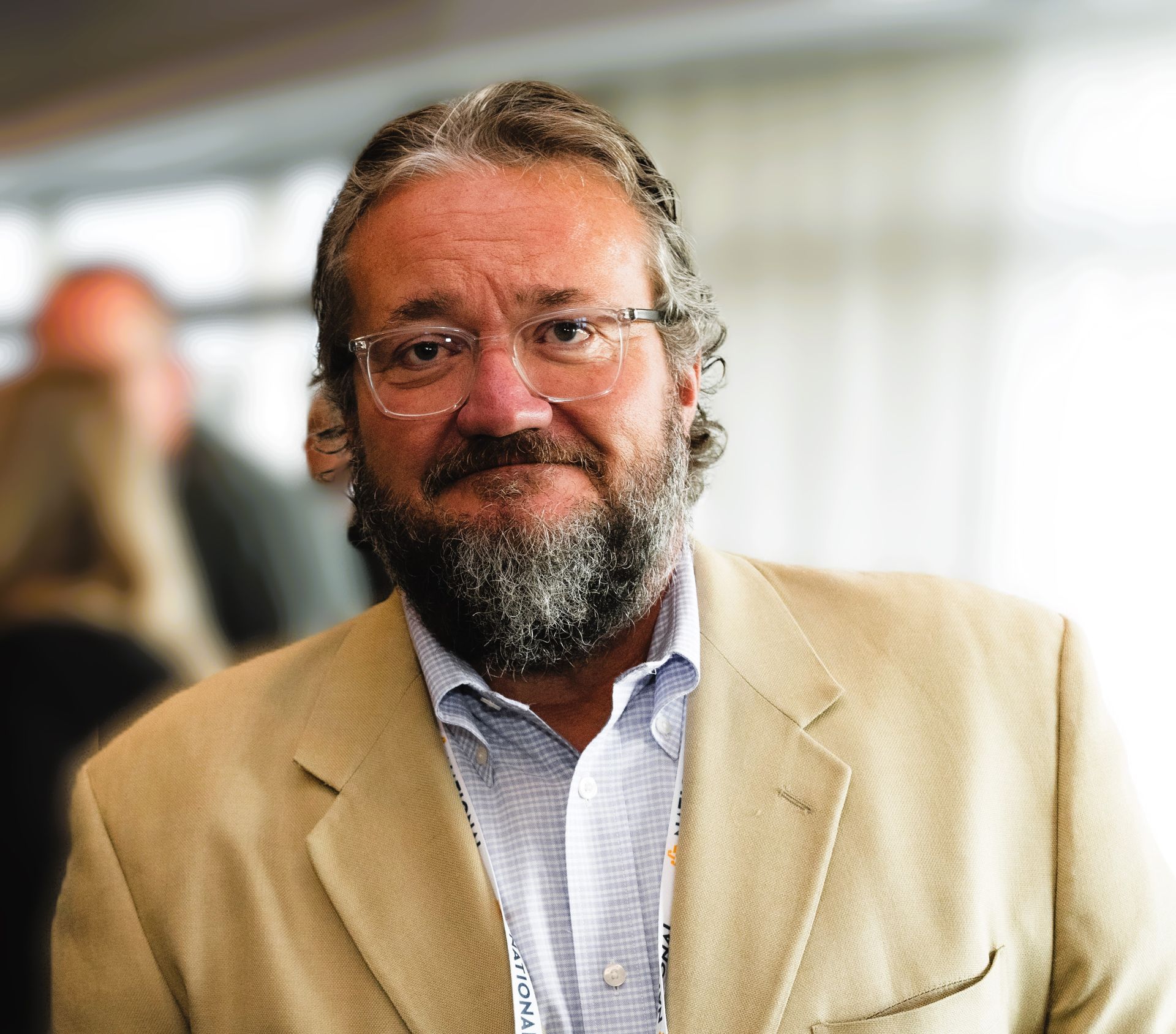 Man in a beige blazer and glasses smiling indoors with a blurred background