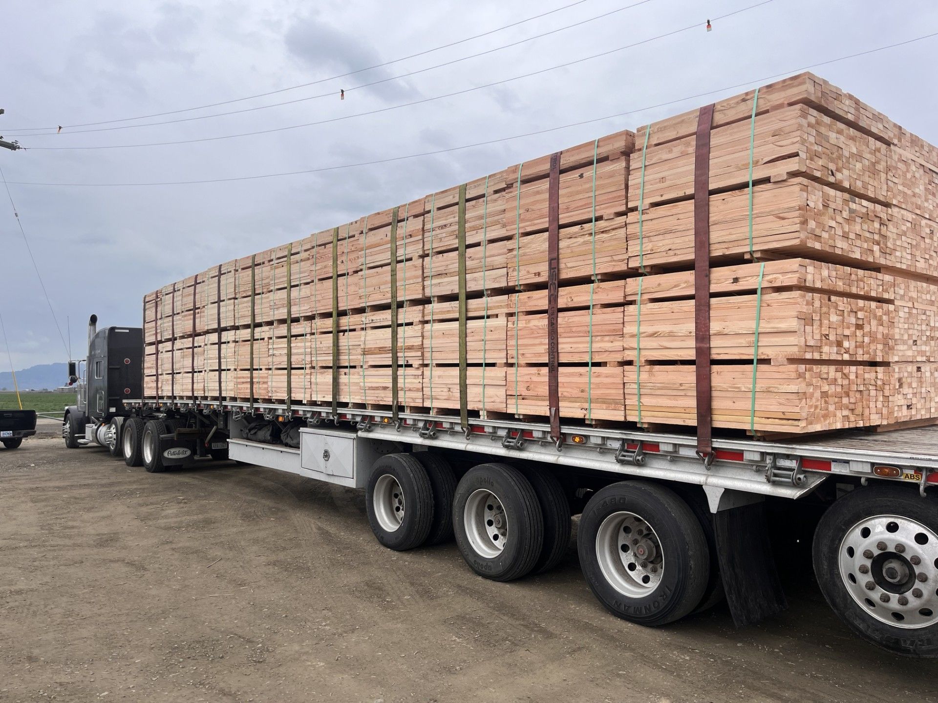 Flatbed semi-truck loaded with stacked lumber in an open yard under a cloudy sky