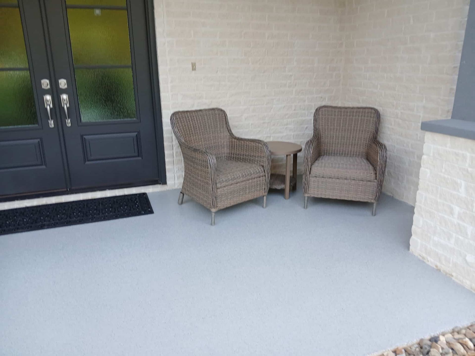 Two wicker chairs and small table on a gray porch, beside black double doors.