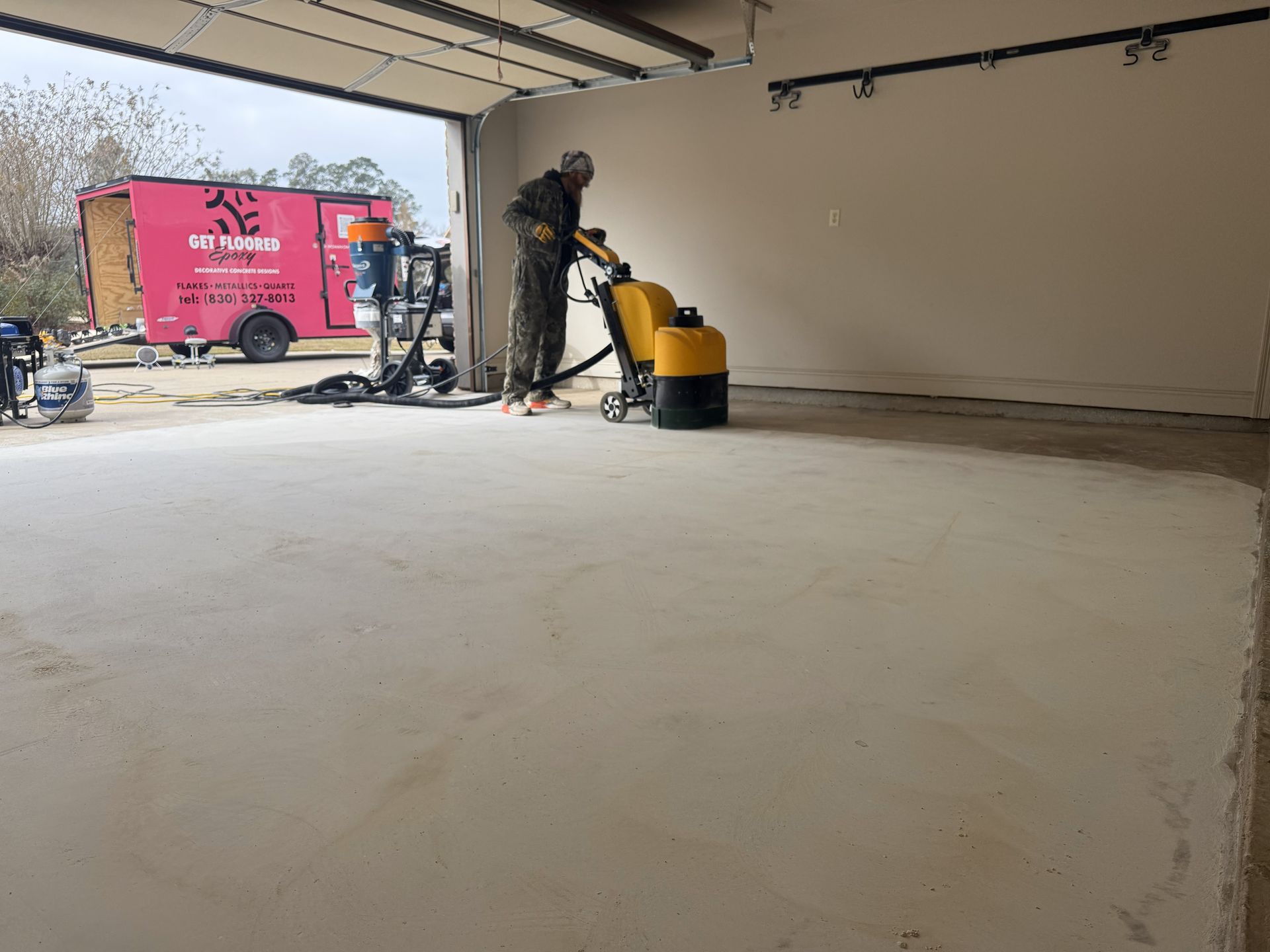 Person grinding a concrete floor inside a garage. A pink trailer is parked outside.