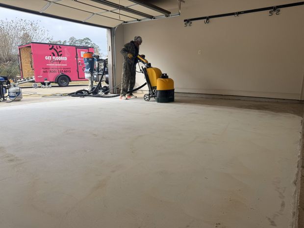 Person operating a yellow concrete floor grinder inside a building with a pink trailer outside.