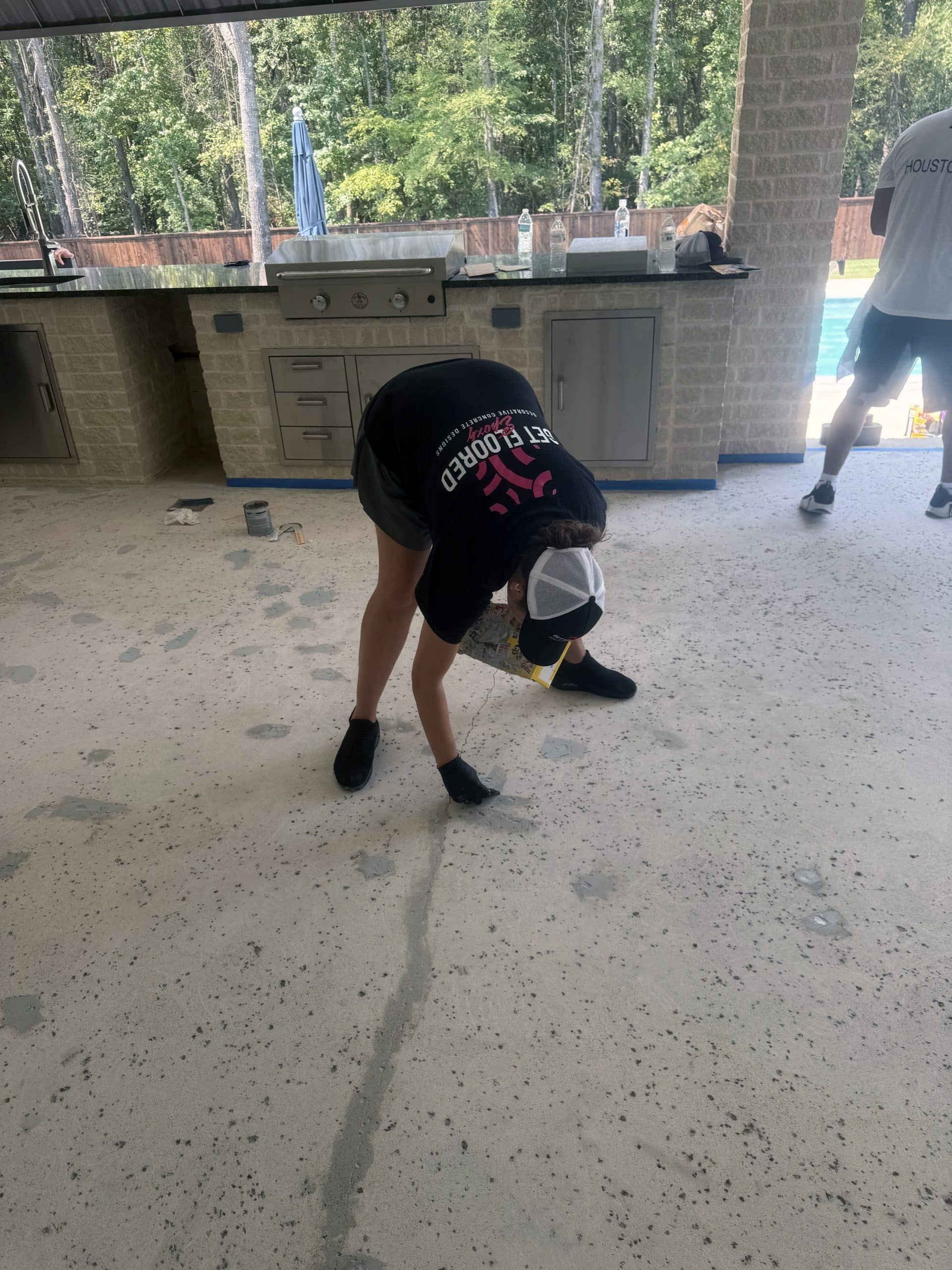 Person bent over, cleaning speckled concrete floor near outdoor kitchen, another person nearby.