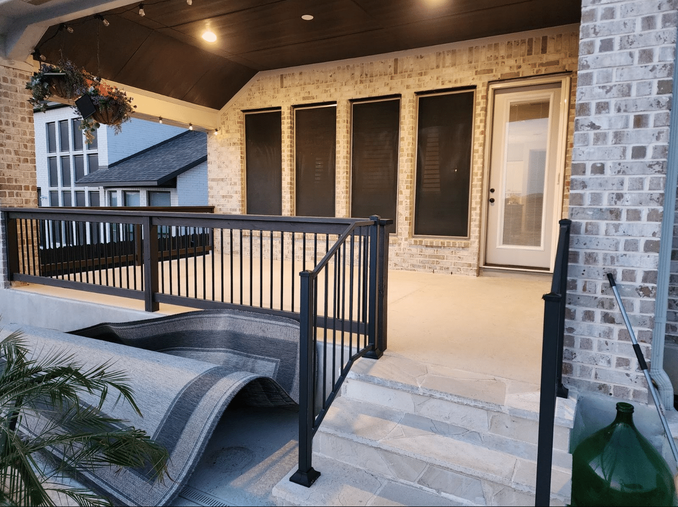 Outdoor covered patio with dark railing, brick walls, and a light-colored floor, with steps leading to a curved structure.