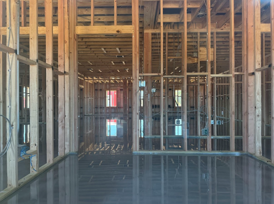 Interior of a building under construction, wooden framing, concrete floor, windows visible, sunlight.