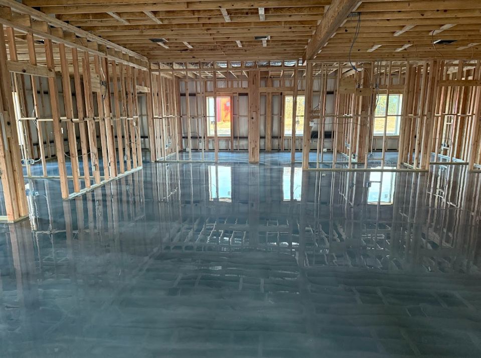 Interior of a building under construction, showing wooden framing and a shiny concrete floor. Windows in the distance.
