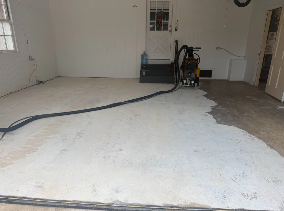 Garage floor being ground down by a machine. The area is partially ground; the walls are white.