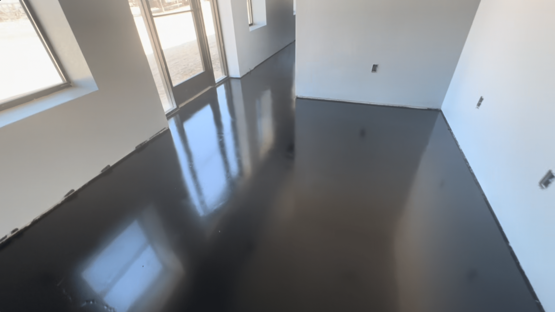 Glossy, dark gray epoxy floor in a room with white walls and windows reflecting light.