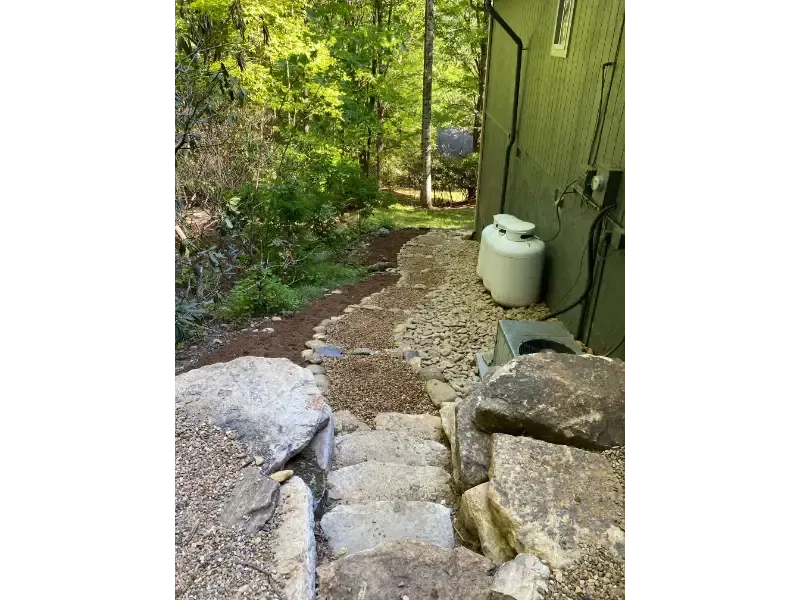 Rocky outdoor path beside a green building and a white tank, leading into a wooded area.
