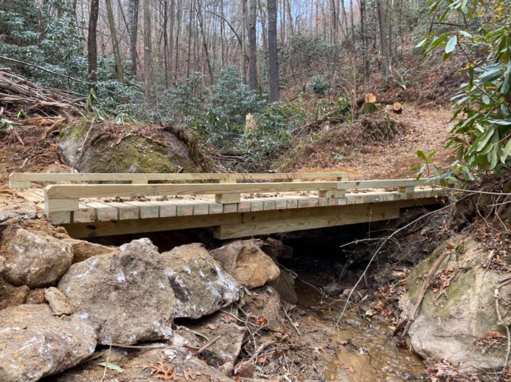 Wooden bridge over a creek in a wooded area, built with rocks on the creek bank.