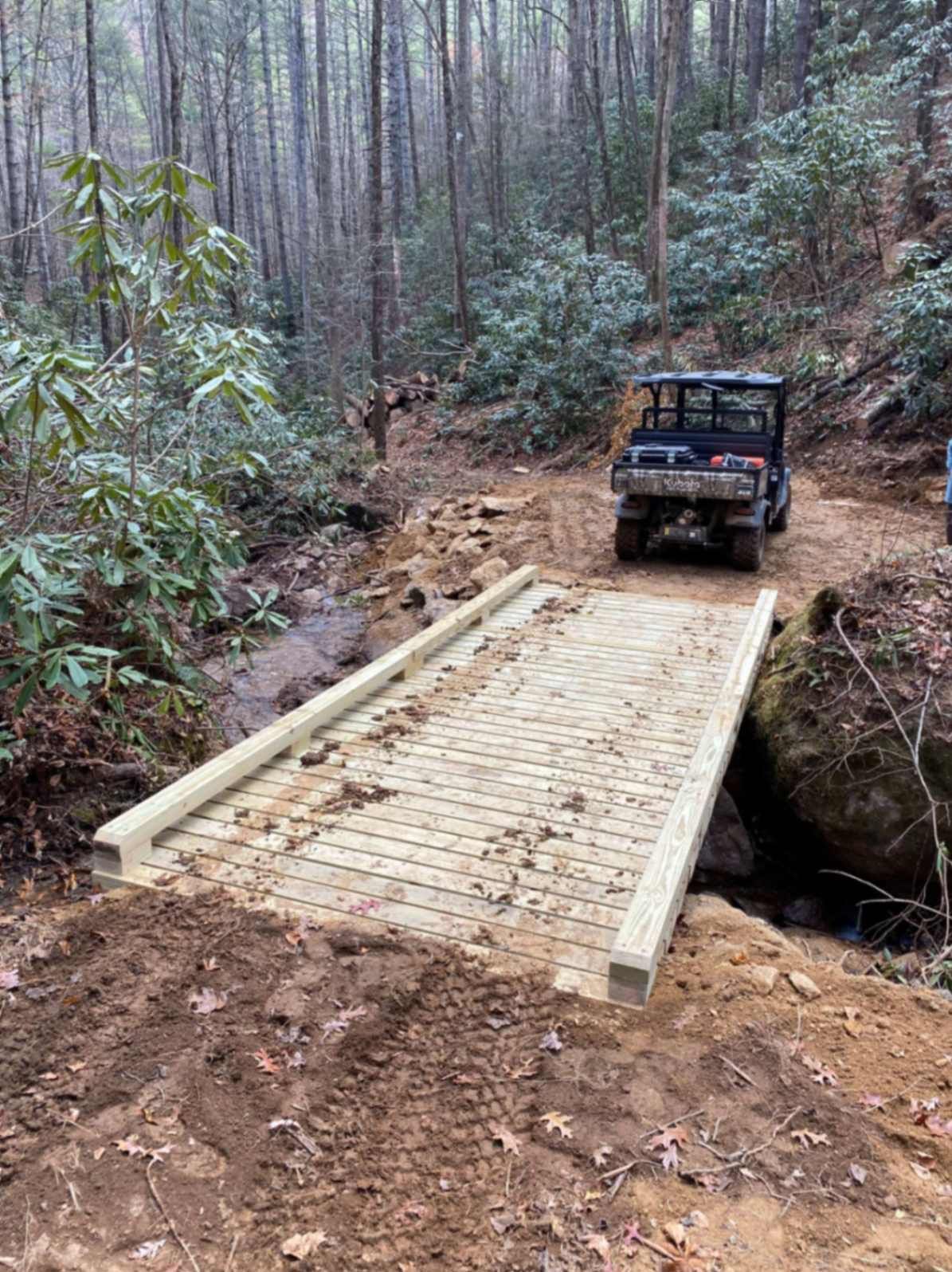 Wooden bridge over a creek with a utility vehicle parked on the path in a forest.