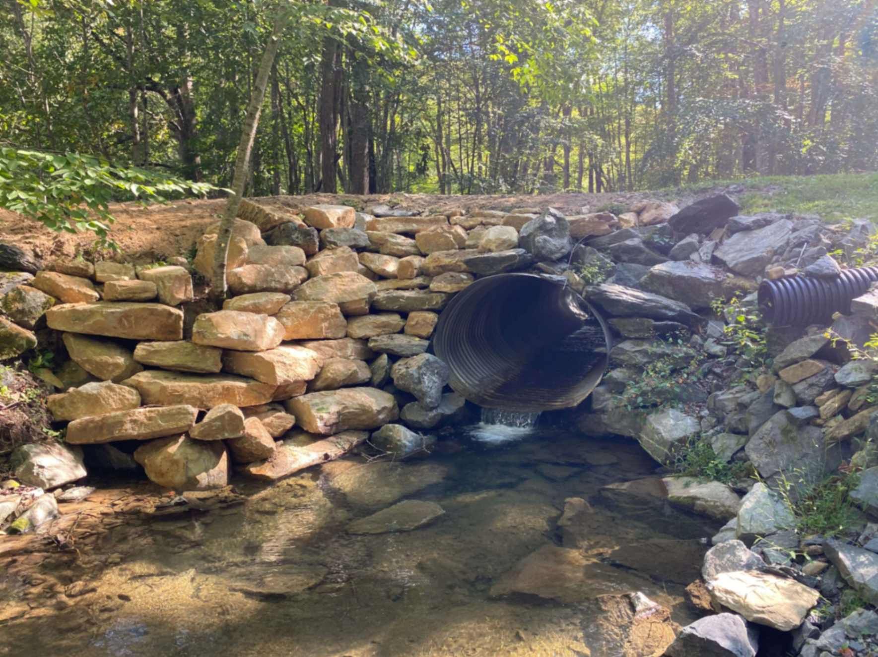 Stone-lined stream with culvert pipe, leading to a wooded area, sunlight filtering through trees.