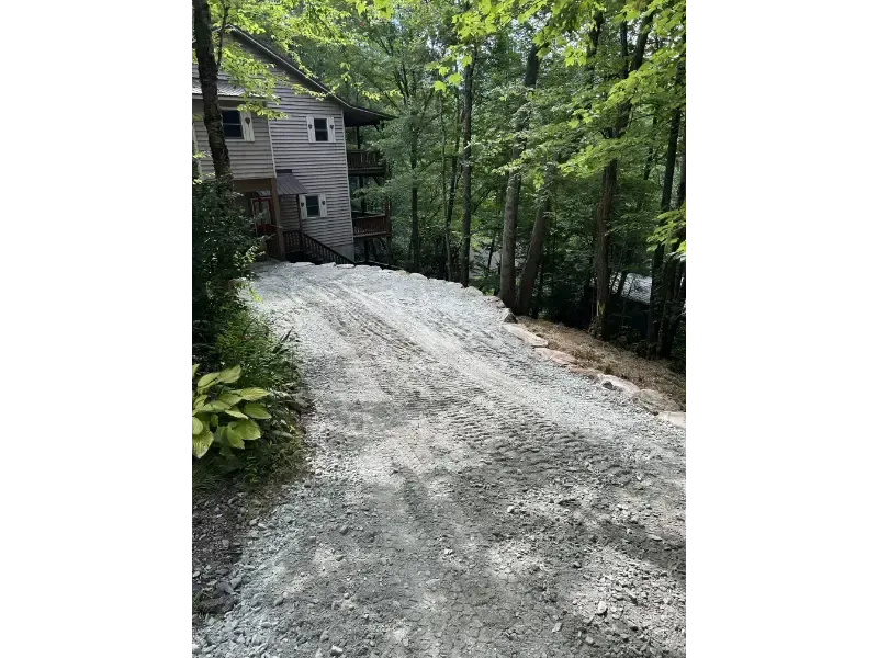 Gravel driveway beside a gray house, surrounded by dense green trees on a wooded slope