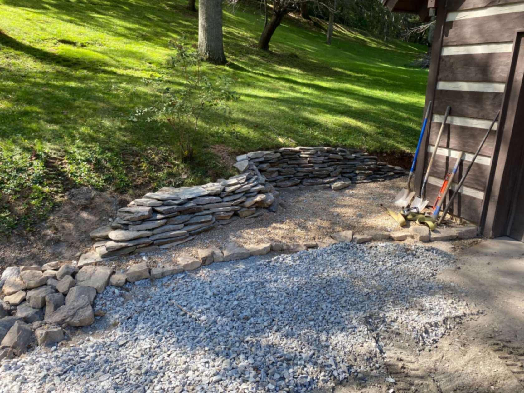 A stone retaining wall under construction next to a log cabin, with gravel and tools on the ground.