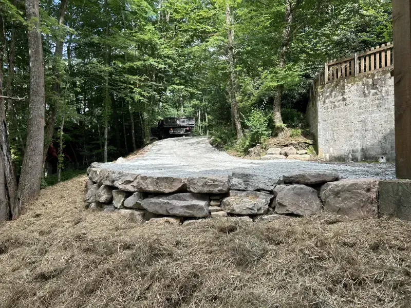 Stone driveway ramp in a wooded area beside a retaining wall and parked car