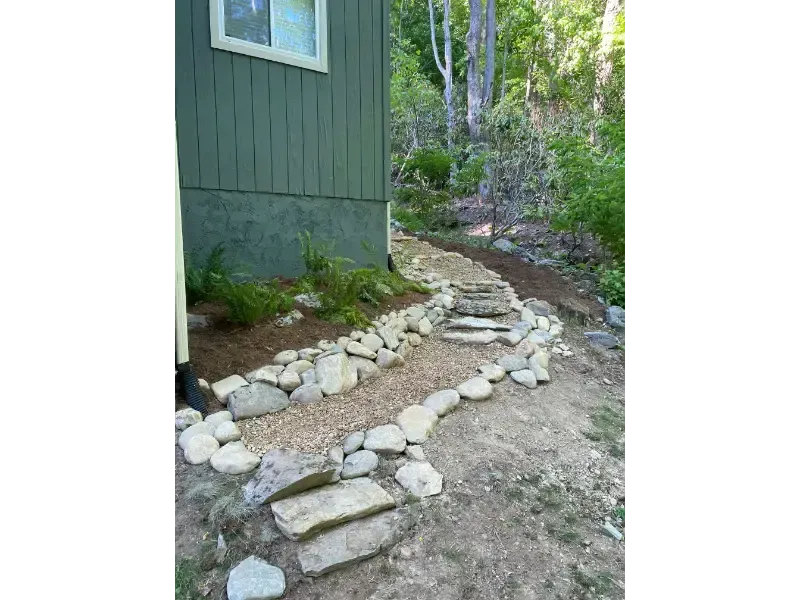 Rock-lined garden path beside a green house, leading into a wooded area.