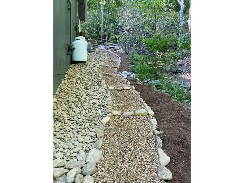 Gravel garden path with stone edging beside a white tank and green shrubs in a wooded area