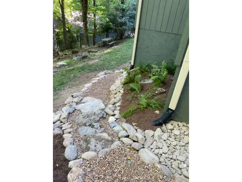 Rock-lined garden path beside a green house leading into a wooded yard