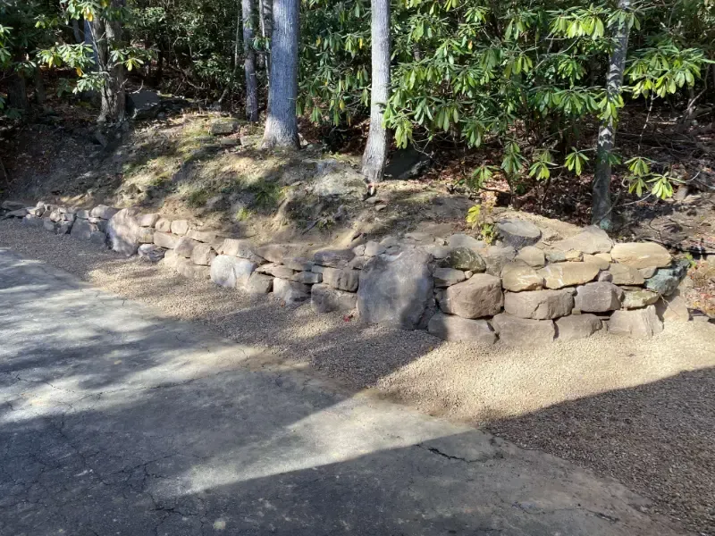 Stone retaining wall beside a dirt path in a wooded area, with sunlight and tree shadows.