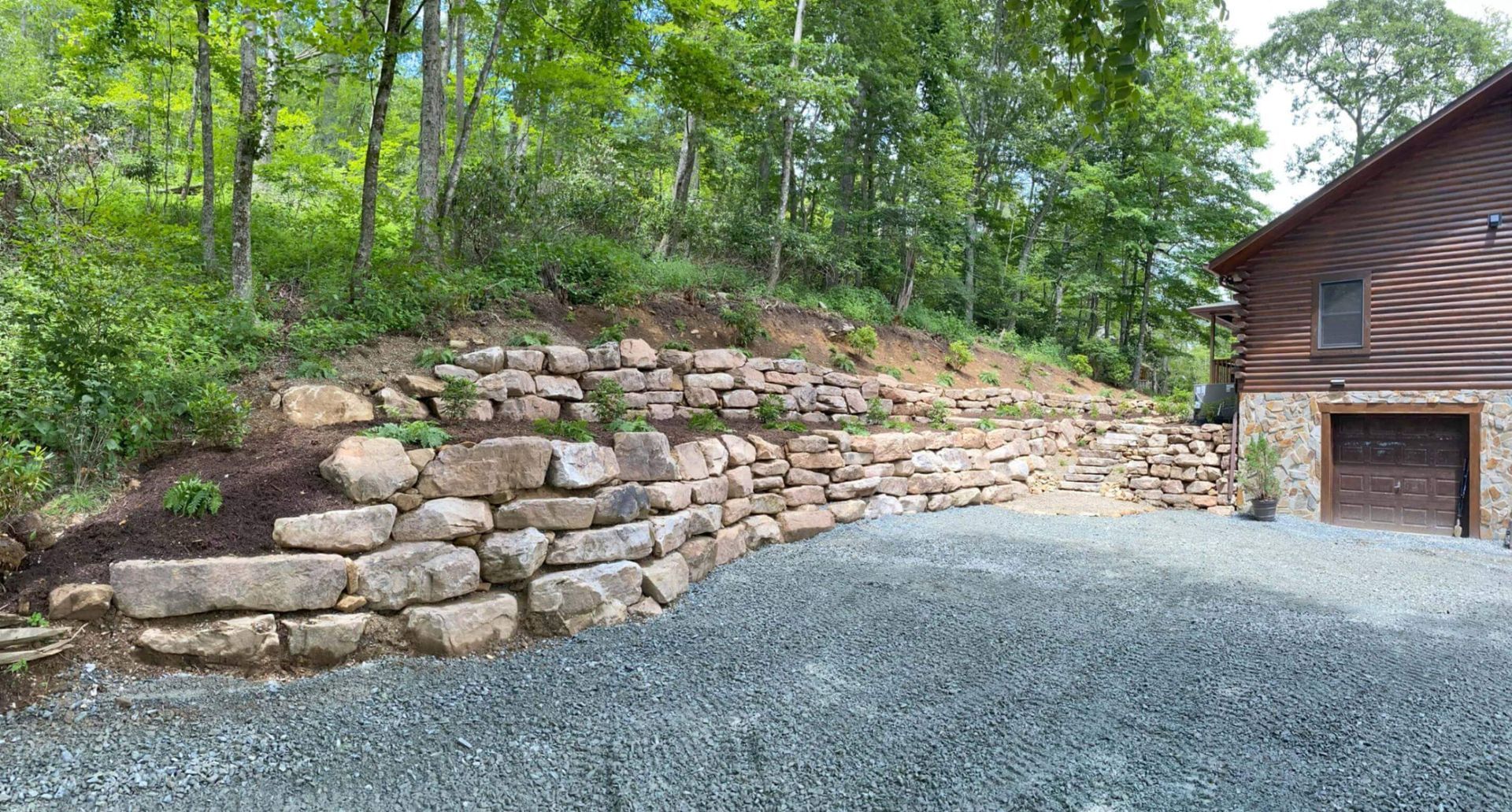 Wooded driveway beside a stone retaining wall and rustic cabin with gravel foreground
