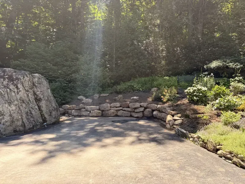 Stone retaining wall and landscaped garden beside a sunlit concrete patio, with trees and shrubs in the background