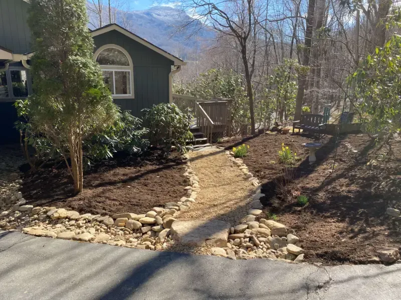 Curved garden path with stone edging leading to a small green house in a wooded yard with mountains beyond