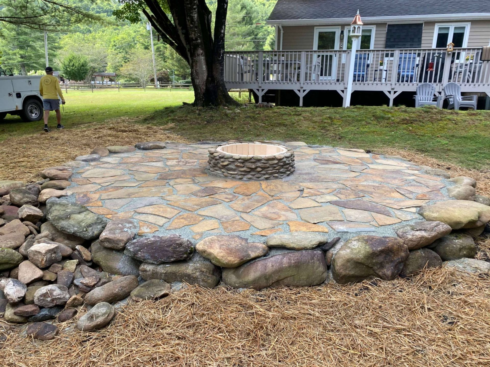 A circular stone fire pit patio built on an earthen slope, surrounded by loose stones and mulch, with a house in the back.