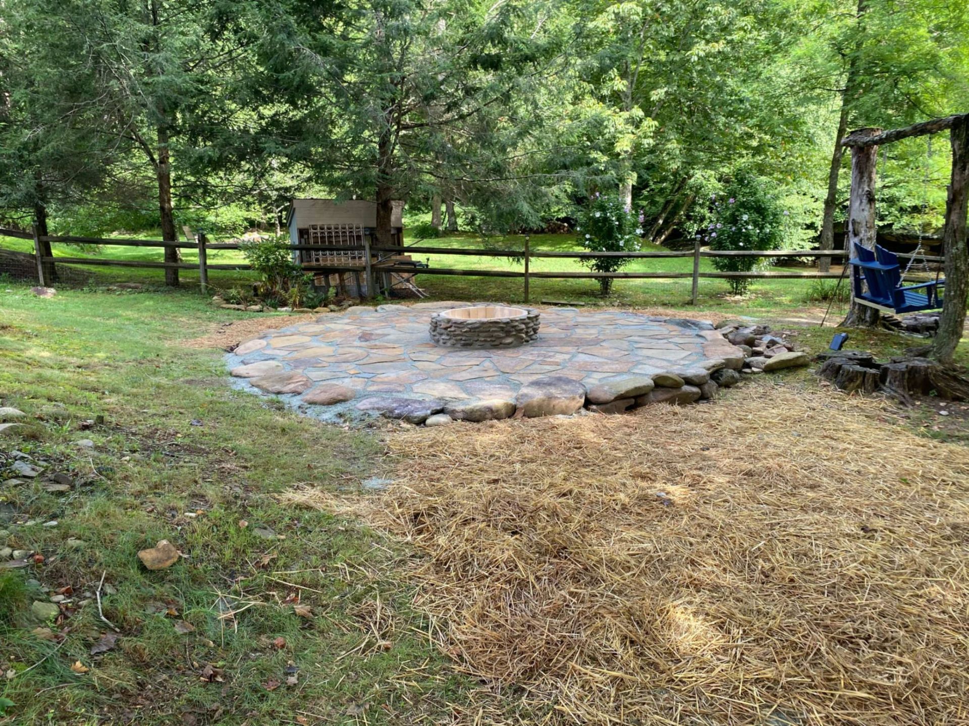Backyard fire pit area with stone circle, log bench, wooden fence, and trees in a grassy yard