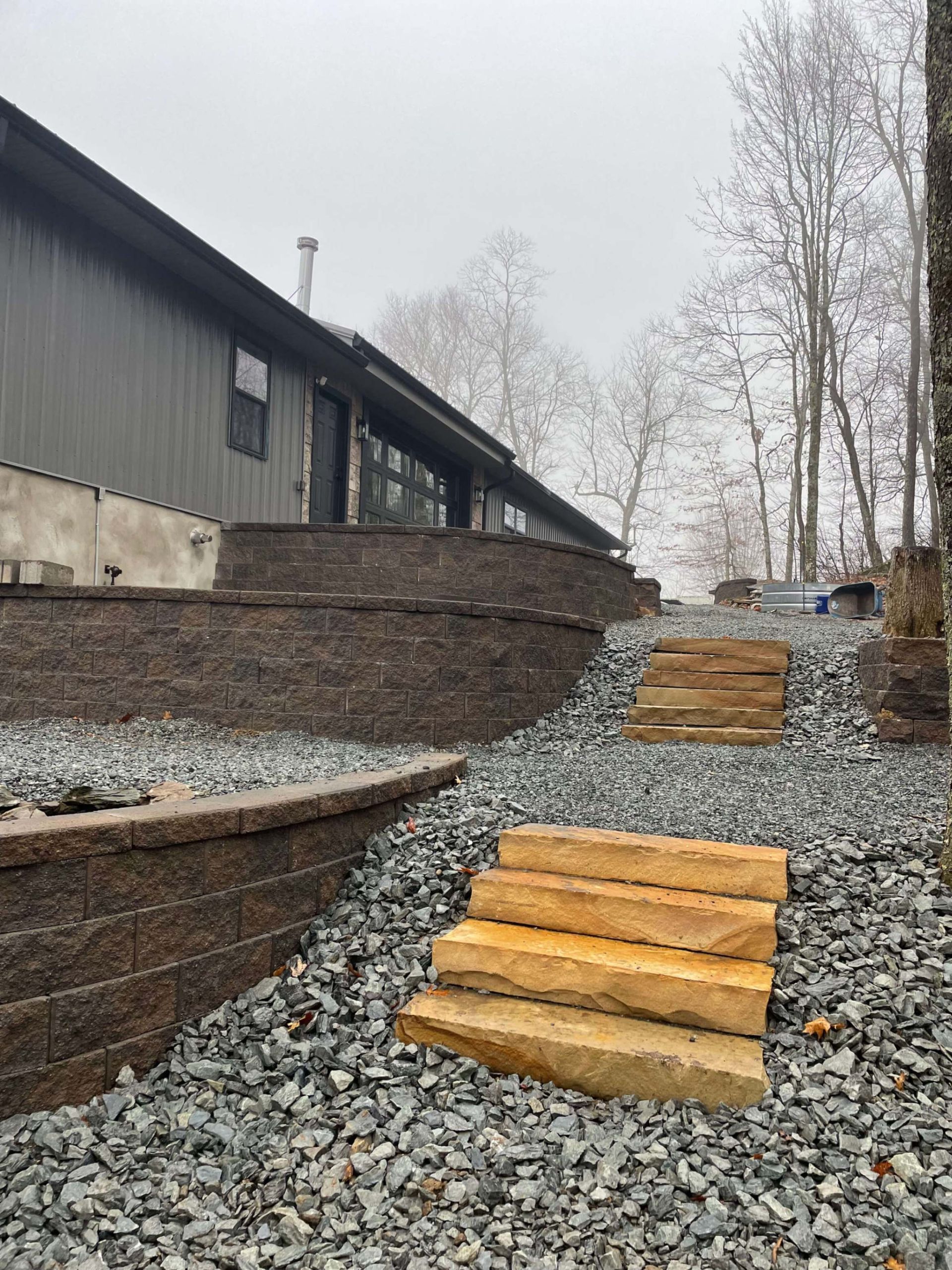 Stone steps lead up a gravel path beside a brown retaining wall and house. Overcast sky.