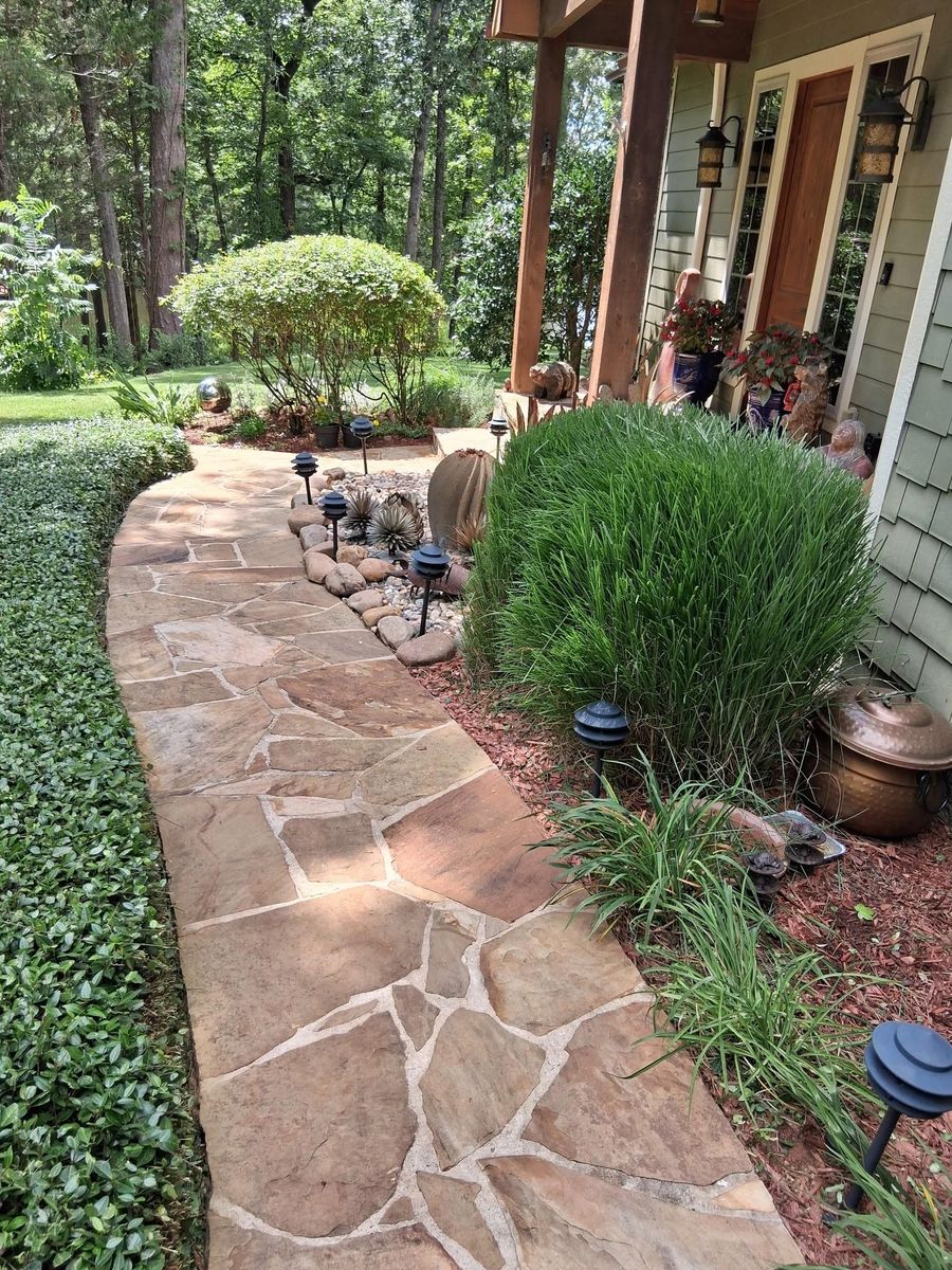 Stone walkway beside a porch, lined with hedges and potted plants in a wooded garden.