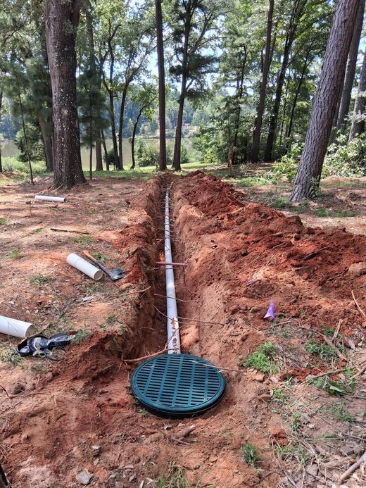 Trench with white pipe and green drain cover in a wooded yard, with exposed red soil and construction debris