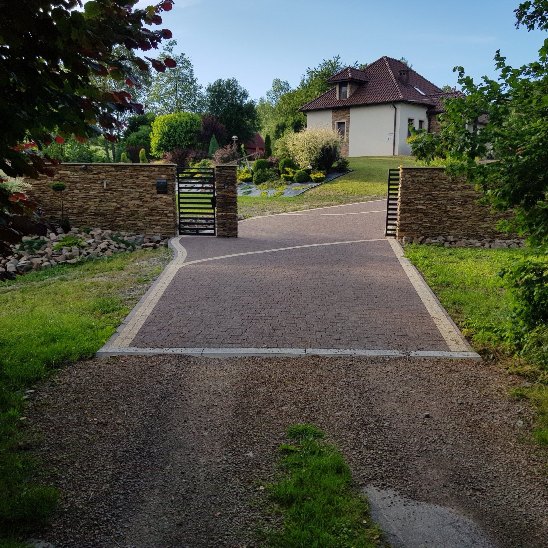Driveway with black gate, stone walls, leading to a house with a red-tiled roof, surrounded by green trees and grass.